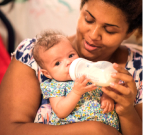 Photo of a woman holding a baby with one arm and a bottle to its mouth with another. The baby has both hands on the bottle