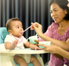 Photo of a baby sitting in a high chair. A woman is sitting next to the baby holding a cup, and another pair of hands is feeding the baby with a spoon.