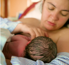 Photo of a woman in a bed with a hospital blanket attempting to get a newborn to latch.