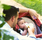 Photo of a man kneeling next to a stroller and holding a bottle to a baby's mouth. The baby has both hands on the bottle and appears to be drinking it.