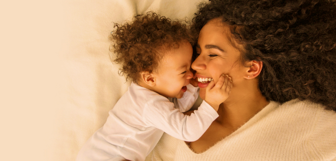 A photo of a dark-haired woman smiling and laying next to a baby. The baby is smily and has both hands on the woman's face.