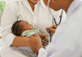 Photo of a woman in a white shirt holding a sleeping baby on her lap. Another person in a white coat is wearing a stethoscope and hold it to the baby.