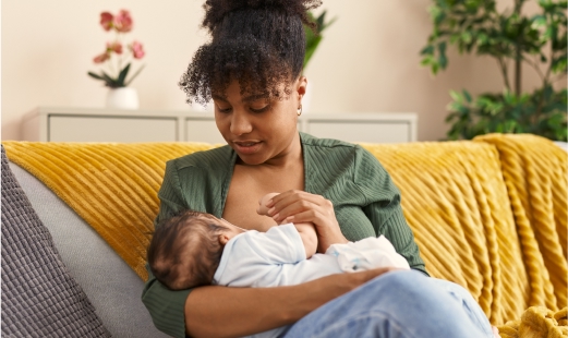 A photo of a dark-haired woman sitting on a sofa, nursing a baby, and holding its hand.